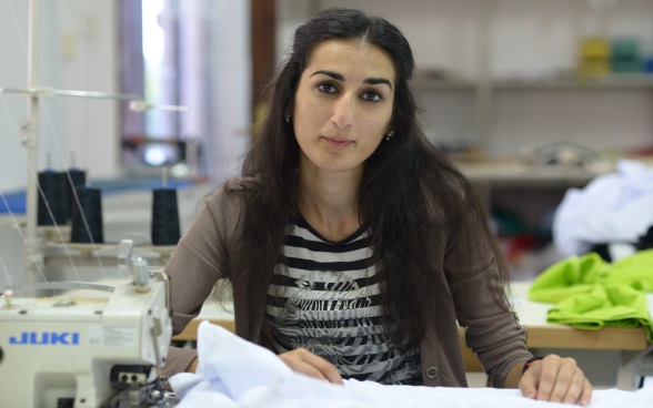  Une jeune femme est assise à côté d’une machine à coudre, les mains posées sur du tissu.