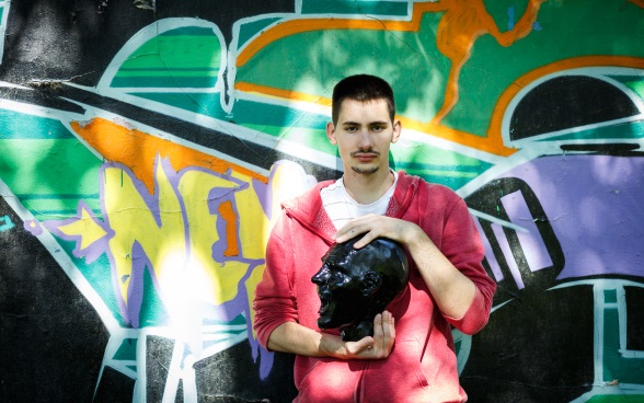  Un jeune homme tenant dans les mains une tête sculptée pose devant un mur couvert de graffitis.