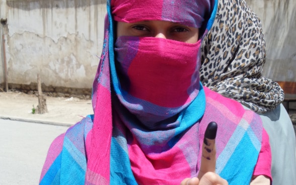 A woman in Afghanistan wearing a colourful veil holds up her left index finger, which has black ink on it after casting her vote.