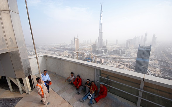 Bauarbeiter machen eine Pause auf der Terrasse eines Gebäudes in Dubai. Im Hintergrund sind im Dunst zahlreiche Hochhäuser und das höchste Gebäude der Welt, der Burj Khalifa, zu erkennen. © Piotr Zarobkiewicz