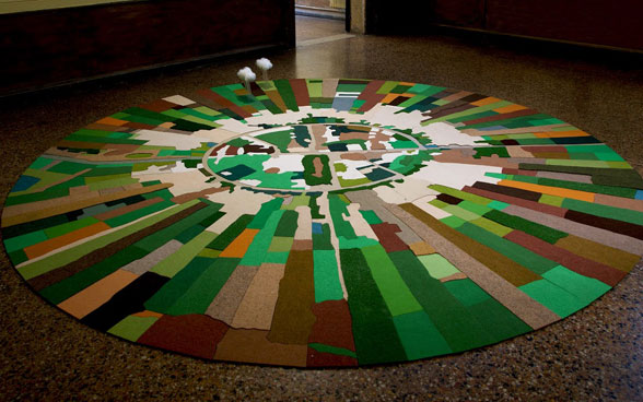 Ein runder Teppich, gefertigt aus verschiedenen Stücken in braun und grün Tönen liegt auf dem Boden. 