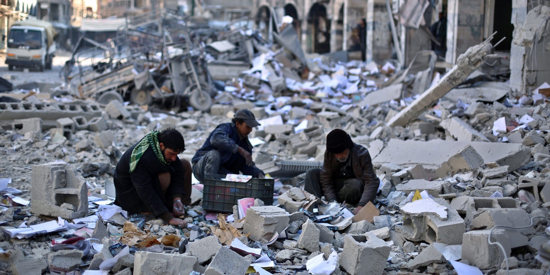 Drei Männer wühlen in den weit verstreuten, das ganze Bild einnehmenden zerbombten Überresten eines Gebäudes, das bei einem Luftangriff getroffen wurde. 