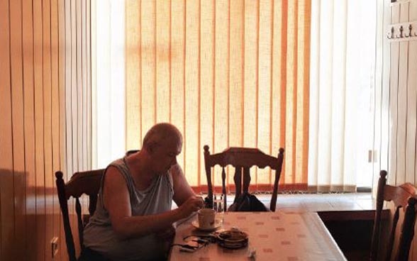  Un anziano siede al tavolo di una stanza in penombra.