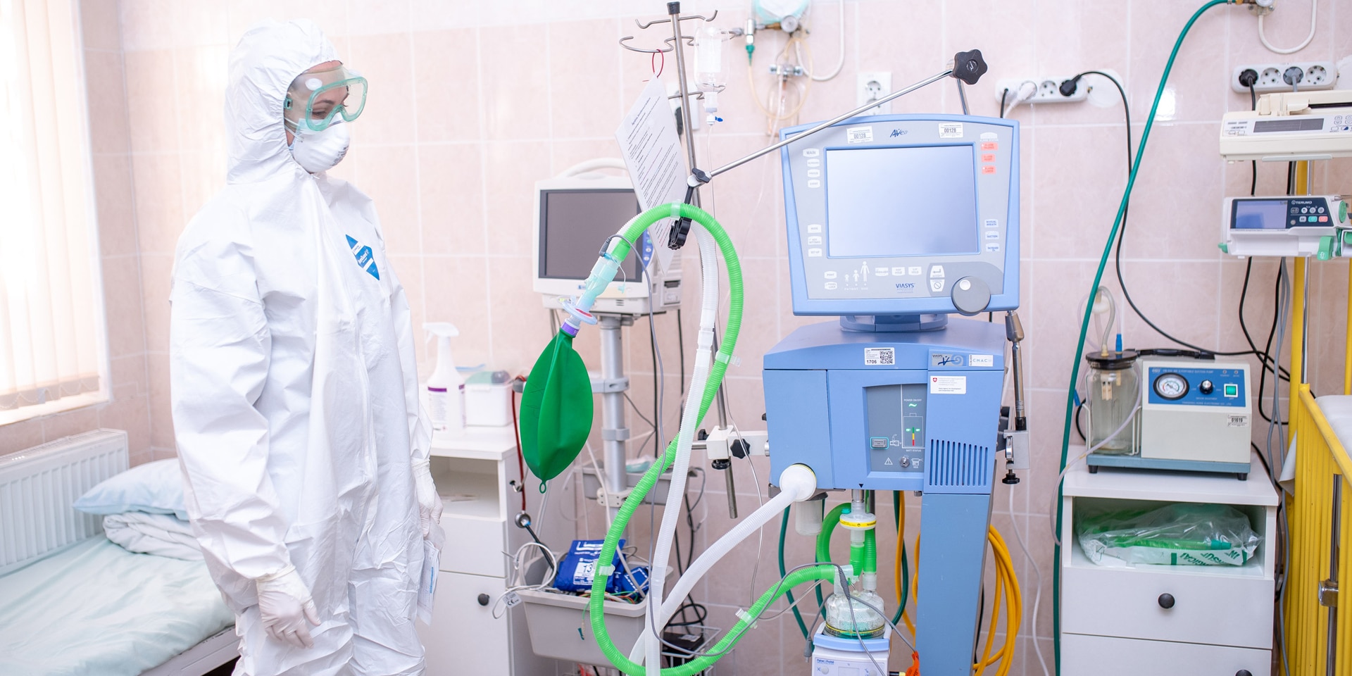 A woman in a hospital room wearing a protective suit, goggles and mask, standing next to hospital equipment. 
