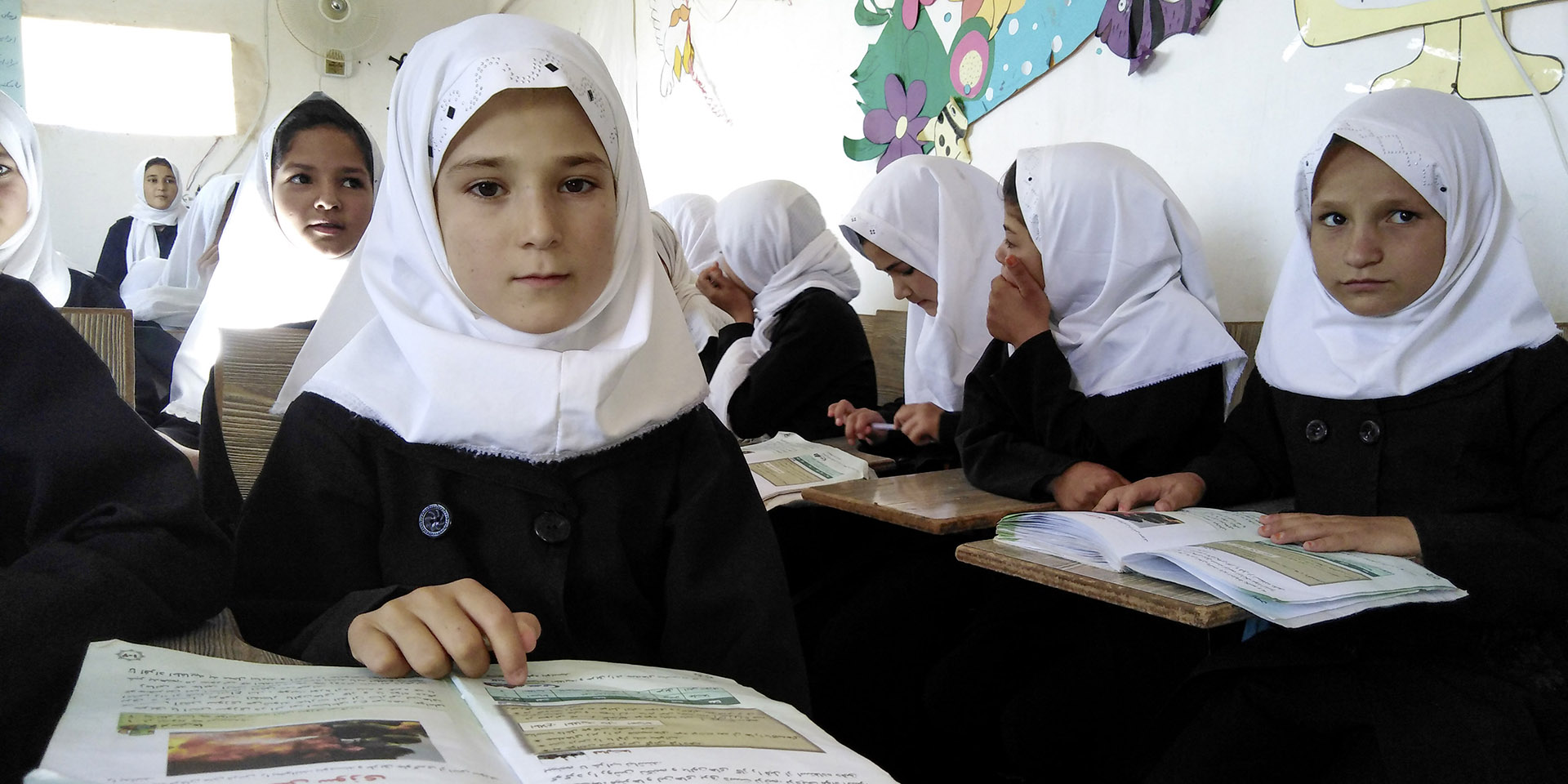 Girls wearing a veil in the foreground. They are in a classroom reading a textbook.
