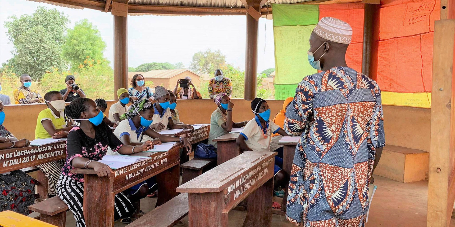 Au Bénin, un enseignant explique devant des élèves entre des pupitres en bois. La salle de classe est couverte d'un toit, soutenu par des poutres en bois.