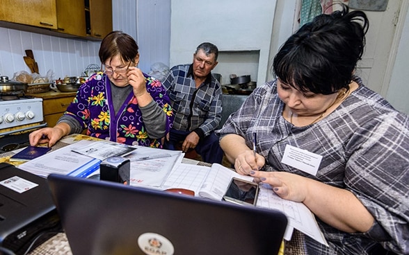 Dos mujeres y un hombre sentados en una cocina, leyendo documentos frente al ordenador. 