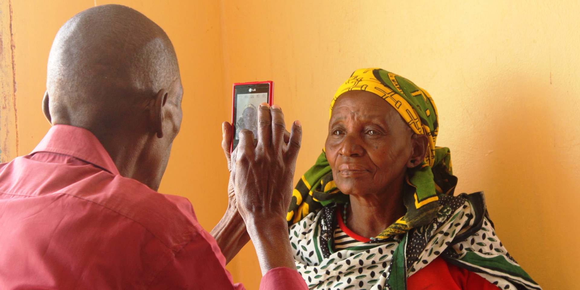 A man takes a portrait photo with his mobile phone of a woman, who is standing in front of an orange wall. 