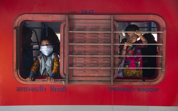 Two children look out of the sliding window of a booth – one with a mask, one without.