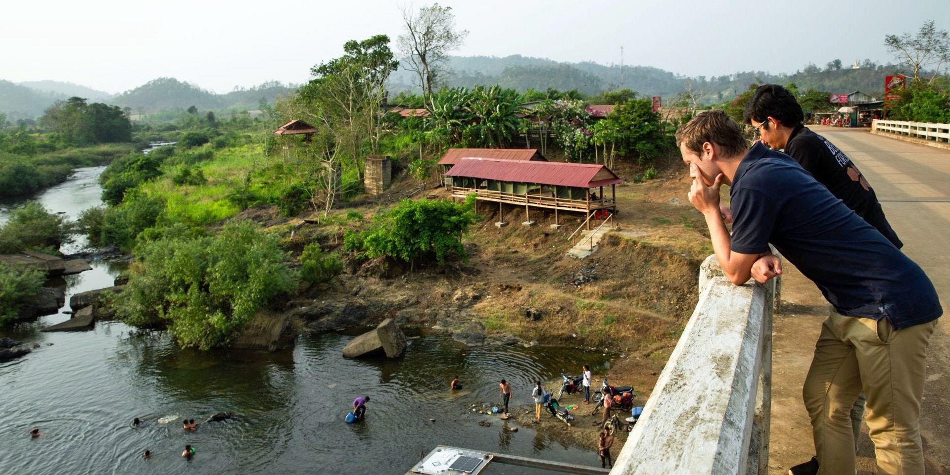  Two people are leaning on the side of a bridge, with people below them working to set up a sensor in the river.