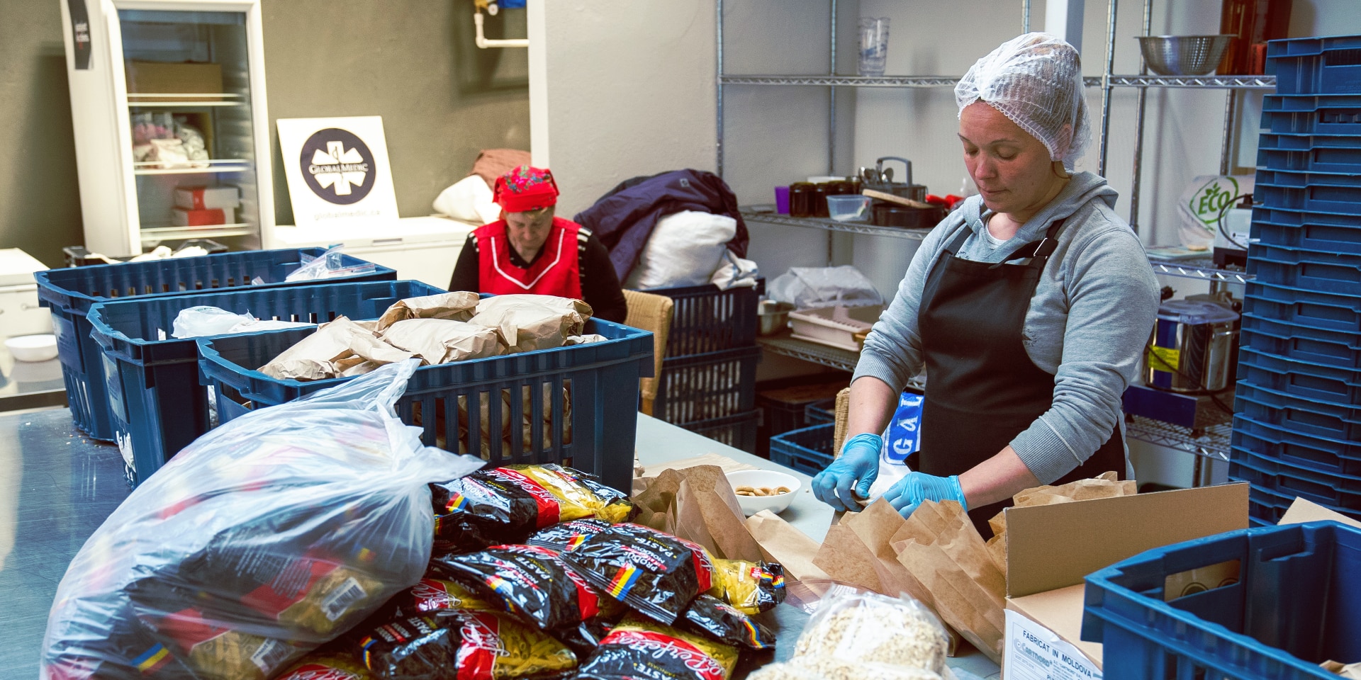 Dos mujeres están empacando alimentos. En una mesa frente a ellos hay muchos paquetes de pasta.