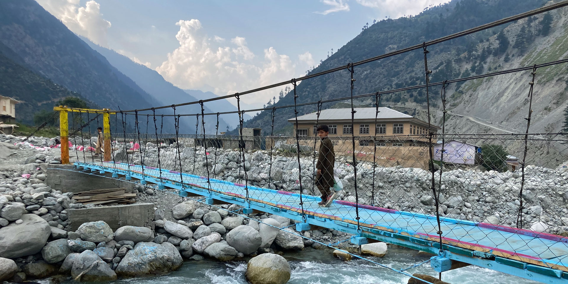 Ein Junge überquert eine Seilbrücke in der Nähe von Mankiala, Pakistan. Eine Fläche aus großen Steinen säumt einen Bach.