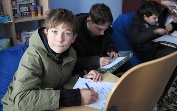 Ein Junge sitzt in einem Raum und schreibt auf ein Blatt Papier, das auf dem Stuhl vor ihm liegt. Um ihn herum lernen andere Kinder in einem ähnlichen Setup.