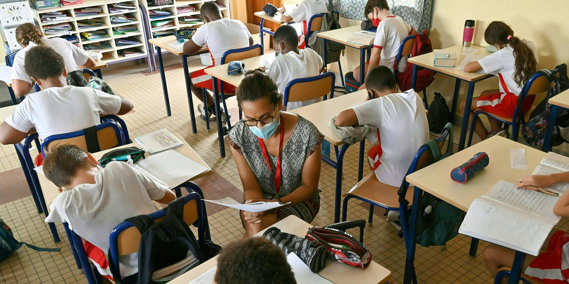Children are sitting between school desks. The teacher helps them with their schoolwork.