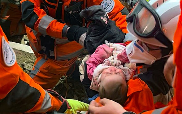 A baby lies in the arms of a woman wearing a helmet and orange clothes, around her two other members of the rescue chain.
