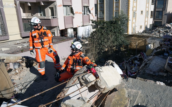 Deux hommes vêtus d'orange se tiennent sur les décombres d'une maison qui s'est effondrée.