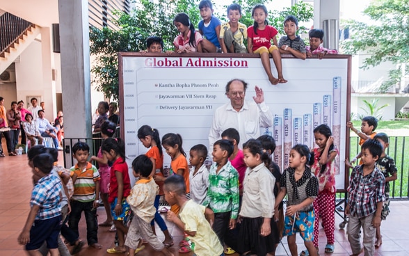 Beat Richner rodeado de niños en Camboya en 2013. Detrás de él, un tablón con un gráfico muestra cuántos pequeños pacientes se atienden cada año en los hospitales.