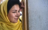 A smiling Afghan woman wearing a traditional yellow headscarf leans on a door frame.