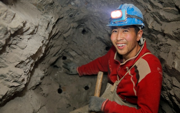 Un uomo, seduto in una miniera, con indosso un maglione rosso e un casco da minatore con torcia. 