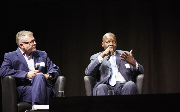 Mansour N’Diaye siede sul palco e parla al microfono. Dominique Guenat gli siede di fianco e ascolta con attenzione