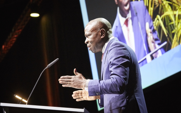 Gilbert F. Houngbo steht auf der Bühne vor dem Rednerpult. Im Hintergrund sieht man eine Leinwand, auf der die Rede übertragen wird.