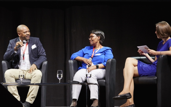 Malijeng Ngqaleni and Melanie Pfändler sitting on stage listening to Sithole Mbanga, who is speaking into a microphone. 