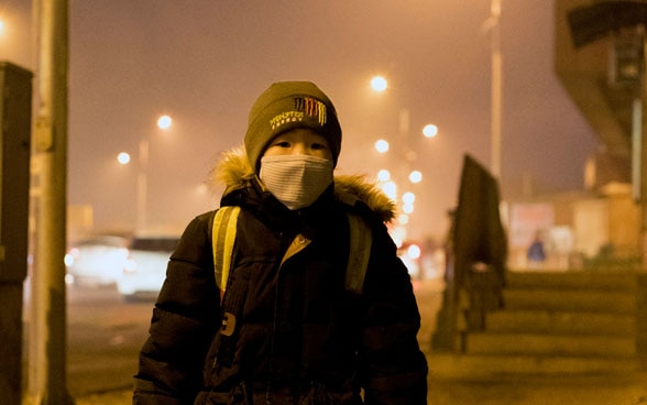 A boy with a school rucksack and face mask stands waiting at a bus stop in Ulaanbaatar.