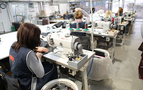 Dans une grande pièce, cinq femmes sont assises l’une derrière l’autre derrière des machines à coudre et cousent.