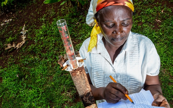 Une jeune agricultrice kenyane nommée Mercy Wambui se tient sur l'herbe. Elle mesure l'eau de pluie et inscrit les données sur sa liste.