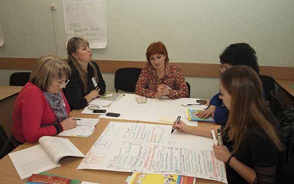 Women holding a discussion around a table covered in notebooks and brochures.