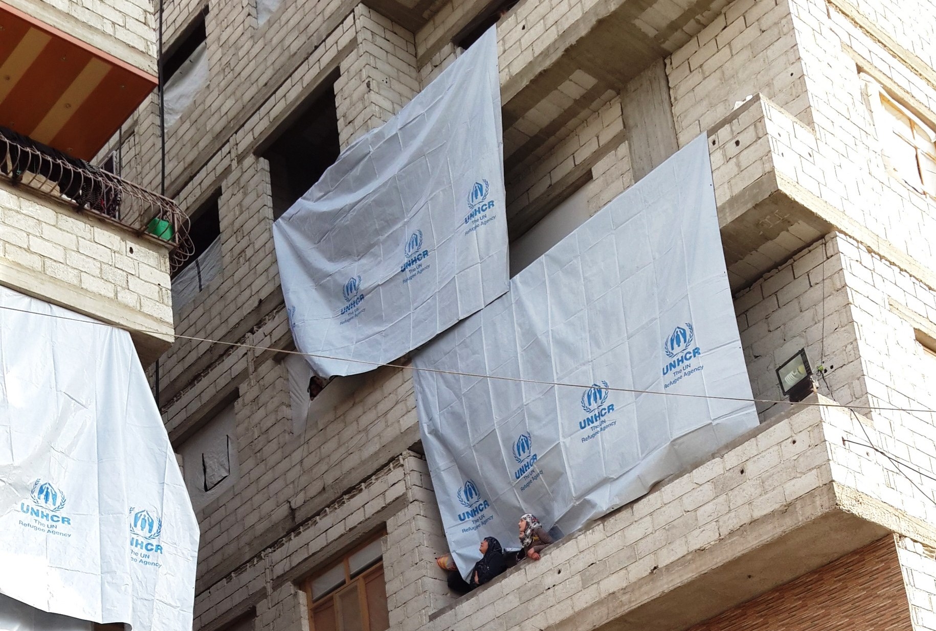 Two women wearing veils on a balcony in a building draped with UNHCR flags. 