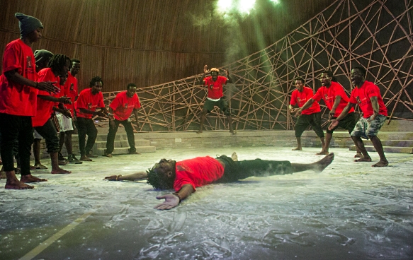 A group of male dancers in red t-shirts and black trousers during a performance at Ubumuntu Arts Festival. A dancer lies motionless in the centre of the stage. Ten other performers dance in a semi-circle around him. 