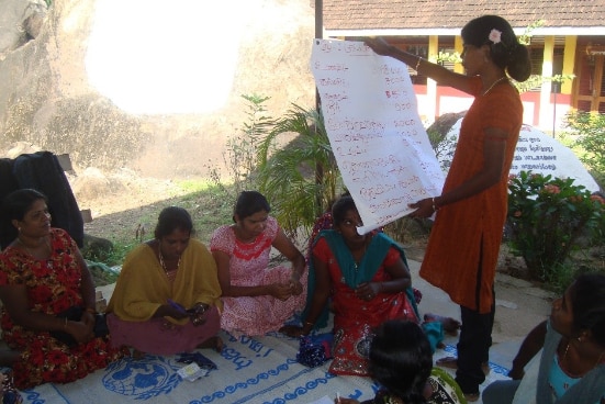 A Sri Lankan woman reading from a large sheet of paper to a group of seated women.