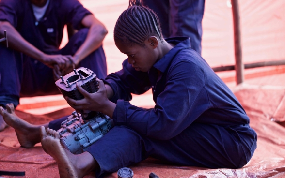 A young woman sits on the ground repairing a motor.