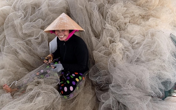 Eine junge Frau sitzt auf einem Haufen Fischernetze. Sie trägt einen Hut und lächelt. Sie hält ein Werkzeug in der Hand und flickt ein Fischernetz. 