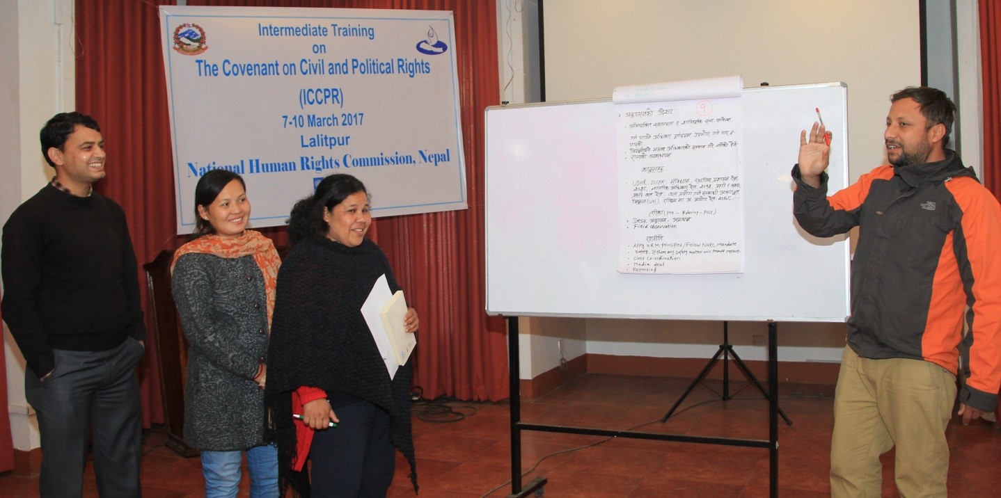 A man stands in front of a whiteboard holding a marker in his raised hand. He is explaining what is written on the board. On the other side of the board stand two women and a man. They listen to their colleague and smile.