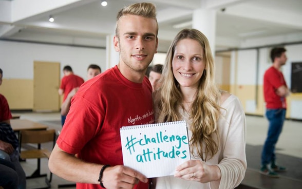 Eine junge Frau und ein junger Mann halten ein Blatt mit der Aufschrift «challenge attitudes» in der Hand.