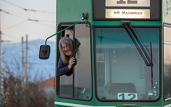 Un hombre con barba se asoma desde la cabina de conducción de un antiguo tranvía de Basilea en una ciudad de Europa del Este.