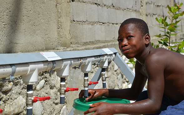un ragazzo beve dell’acqua e riempie una tanica dai rubinetti installati dall’aiuto umanitario della DSC.