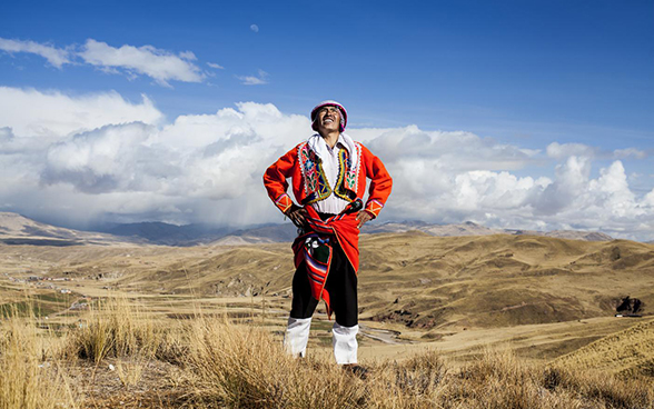 A man in traditional Peruvian clothing stands in open countryside in the Peruvian highlands, and looks up at the sky.