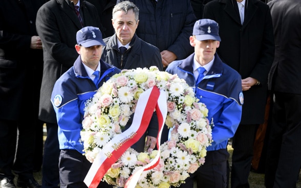 Bundespräsident Cassis steht hinter Blumenkranz, der von zwei Polizisten getragen wird.