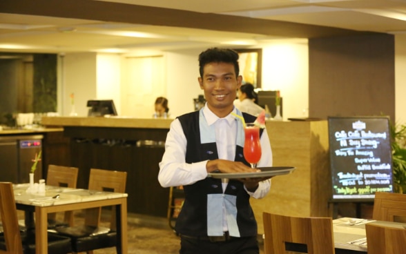 Un jeune serveur souriant dans un restaurant tient entre les mains un plateau sur lequel est posé un cocktail.
