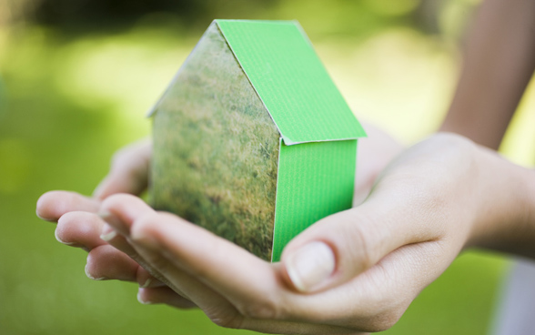 Two hands holding a small green house made of paper.