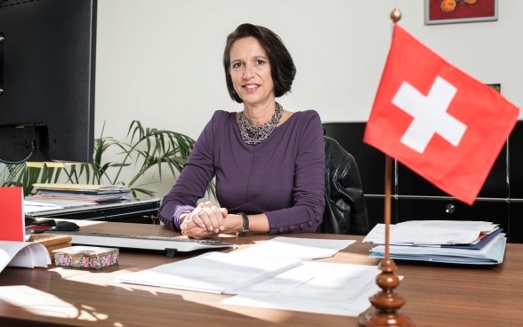 Une femme aux cheveux noirs, Christine Schraner Burgener Burgener, est assise derrière un bureau avec un petit drapeau suisse.