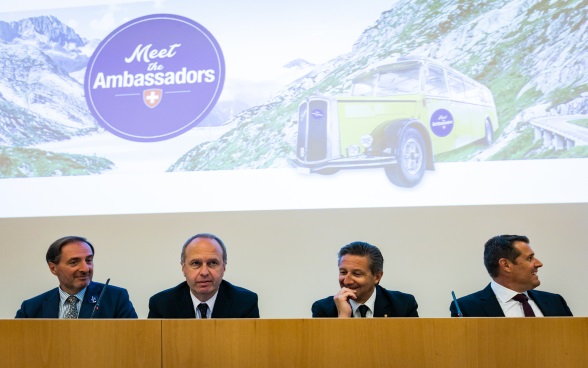 Quattro ambasciatori sul podio dell'auditorium, sullo sfondo una foto della corriera postale Meet the Ambassador