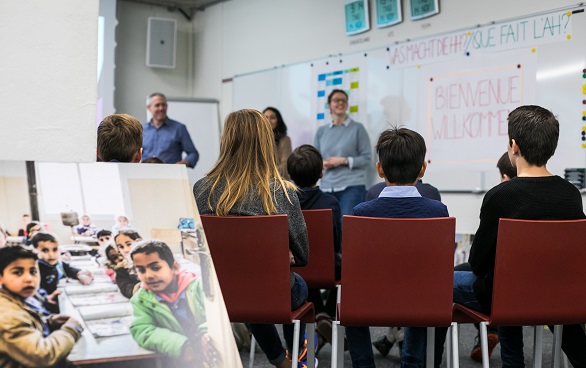 Des enfants écoutent une femme présenter le travail de l'Aide humanitaire de la Suisse.