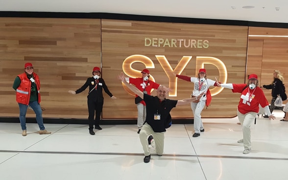 Mitglieder des Teams des Schweizerischen Generalkonsulats in Sydney posieren vor einem Leuchtschild, das die Abflughalle anzeigt. 
