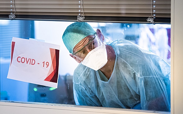  A health worker looks through an intensive care unit window past a taped-on paper sign that says 'COVID–19'. 