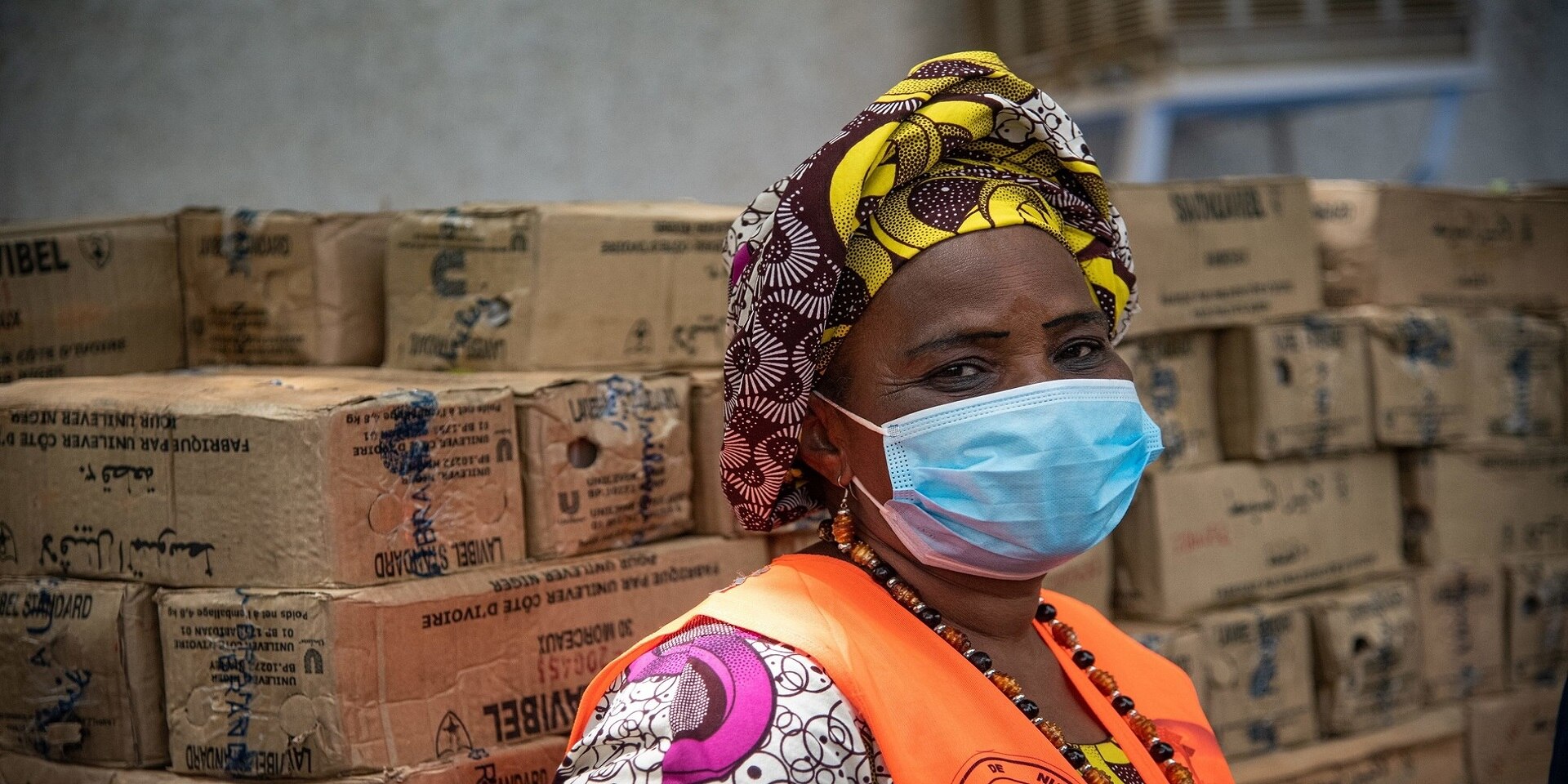 An African woman wearing a mask and an orange vest in a warehouse stacked with boxes