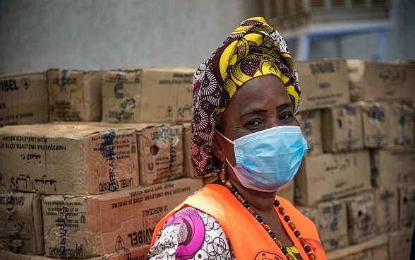An African woman wearing a mask and an orange vest in a warehouse stacked with boxes